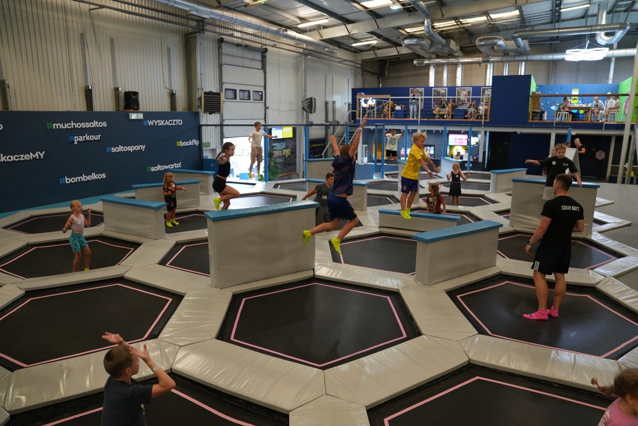 Grupa dzieci skacze na polu trampolin, kilkoro w powietrzu, trener obok